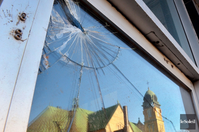 Photo : vue de près d'une partie fracassée d'une fenêtre. Dans le reflet de la fenêtre, on discerne bien un bâtiment ancien de la ville en avant qui ressemble un peu à une église..